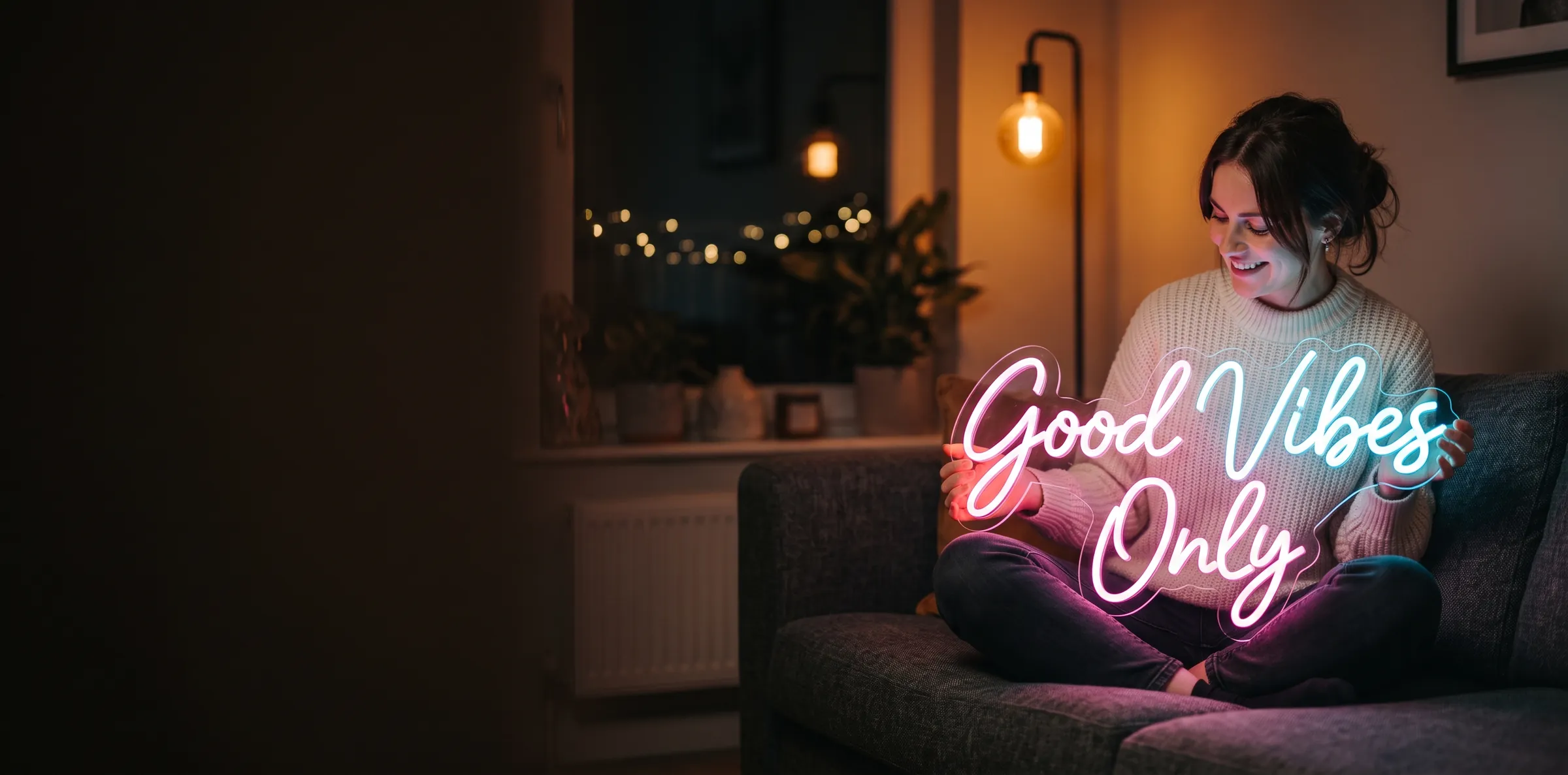 A woman on a sofa in a warm, dimly lit living room smiling at the custom LED neon sign in her lap, reading “Good Vibes Only” in glowing pink and cyan script